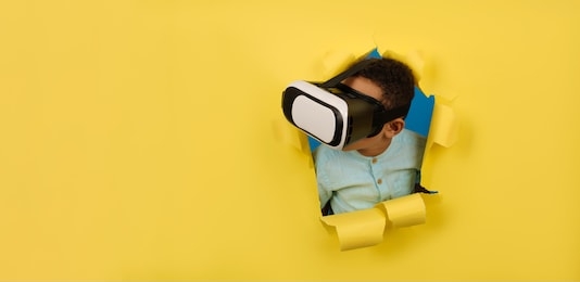 african-american boy in virtual reality vr helmet looks away. child is immersed in world of 3d graphics, considering world of virtual reality in helmet on yellow background torn paper. space for text