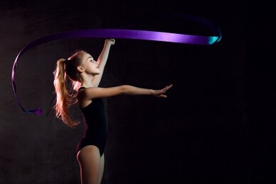 young girl gymnast in black sport body and uppers standing sideways and making exercise with blue gymnastic tape over dark background. rhytmhic gimnastics beauty cocnept
