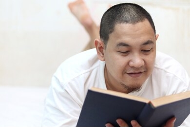 happy male smiling while reading book on bed, leisure and calming activity of smart people enjoy reading before sleep at night