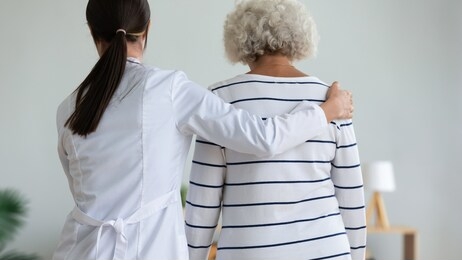 rear back view young caregiver in white coat supporting hugging elderly patient help her to walk, physiotherapist and physiotherapy activity, provide assistance to old woman, homecare nursing concept
