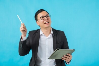 young asian businessman wearing glasses holding tablet and thinking about his business idea isolated on blue background.concept of  success creative startup business.