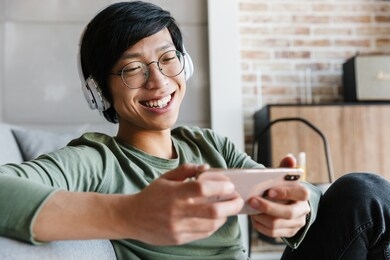 image of handsome young asian man wearing headphones using cellphone while resting in apartment