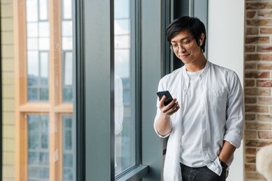 image of handsome young asian man wearing earphones using cellphone while standing by window in office