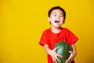 happy portrait asian child or kid cute little boy attractive smile wearing red t-shirt playing holds full watermelon that has not been cut, studio shot isolated on yellow background