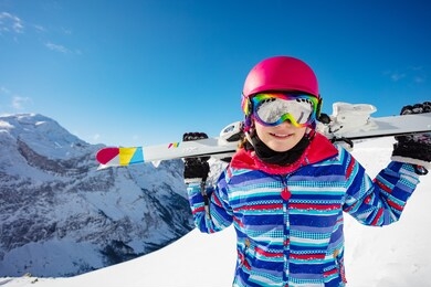 happy cute girl ski on her shoulders smile wearing color glasses and helmet over mountain peaks on background