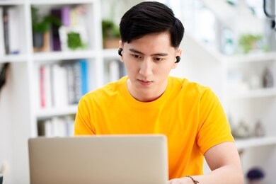 asian young male student use wireless earbuds to have online language courses and searching information via laptop computer at home   