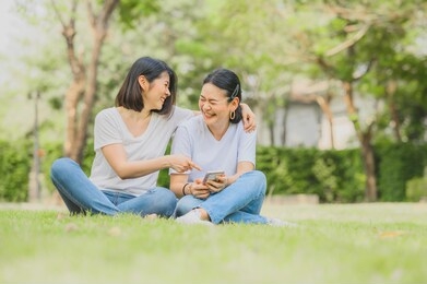 two happy best friends asian women are laughing while using mobile smartphone outdoor in the park.