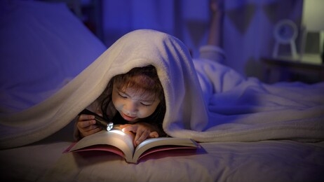 child asian girl reading a book with flashlight under the blanket on bed in a dark bedroom at night before sleeping, comfortable children at home concept