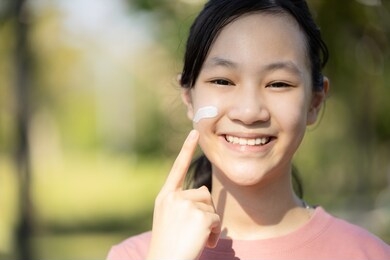 happy smiling asian child girl apply sunscreen lotion on the cheek,protect her face,schoolgirl using skin care product for sensitive skin,sun block day cream on her delicate facial skin in sunny day
