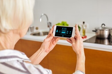 senior woman at home standing at kitchen holding smartphone controlling smart home system back view close-up turning on light