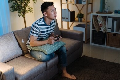 young asian chinese man addicted to video gaming at dark home. concentrated handsome guy in casual wear stay up late house living room. male sitting on sofa holding joystick remote in evening.