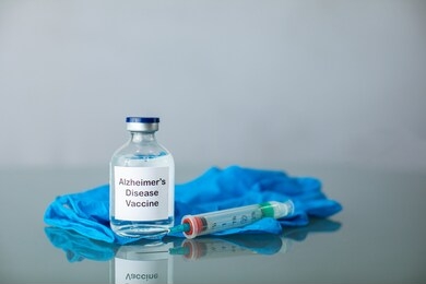a bottle of a clear medicine alzheimer's vaccine along with a syringe and surgical gloves on a plain surface