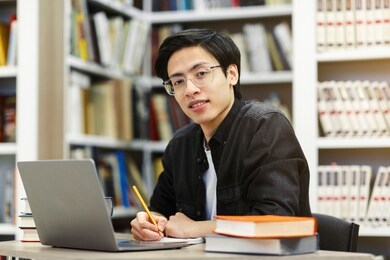 working on research project. portrait of asian male student using laptop at library, writing in notepad, looking at camera