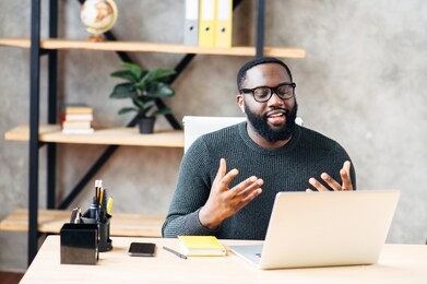 video call. attractive african-american guy is using laptop for video communication, a guy in glasses sits at the table in modern office, looking into webcam and talking online