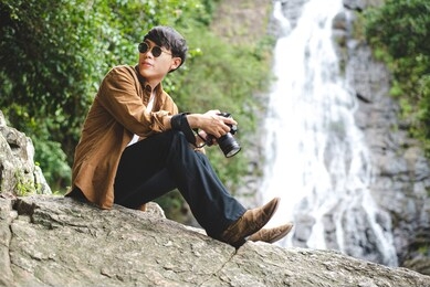 asian man traveler use his camera taking photo of nature with waterfall behide him.