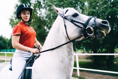 young female equestrian in black helmet and red shirt sitting in saddle of white horse and holding bridle while looking at camera