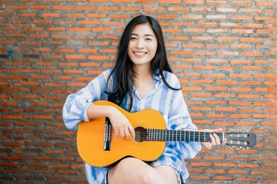beautiful asian female musician performing live in front of audience recording viral video on social media singing playing guitar taking to people, camera vlogging at working at home brick background 