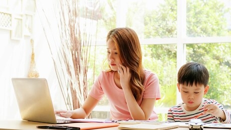 asian mother and son in their living room. mom is busy with remote work while son is drawing something by himself.
