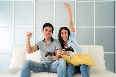 asian couple man ans woman to playing video games with joysticks while sitting in sofa in living room at home, concept of family values and the modern-day game.