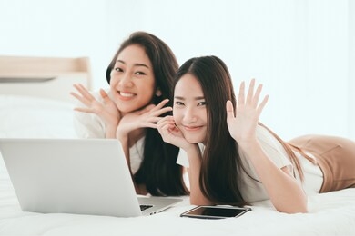 two young asian women spending lazy weekend together lying on bed at home use laptop browse web watching video interesting vlog search best offers via internet buying online.