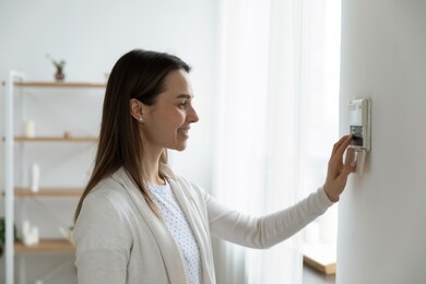 smiling woman standing near wall-mounted device adjusting degrees in living room set comfortable temperature using thermostat home heating system. owner of modern smart house, energy saving concept