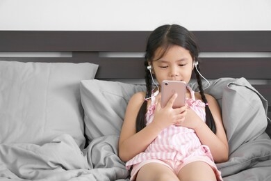 asian child or kid girl smile wear headphones or small talk to learn study online or work from home on smartphone and mobile phone communication or people listen music and leaning relax pillow on bed
