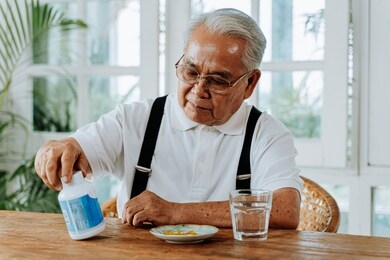 senior asian man with cup of water and pill taking remedy in cozy room at home. old male taking medicine