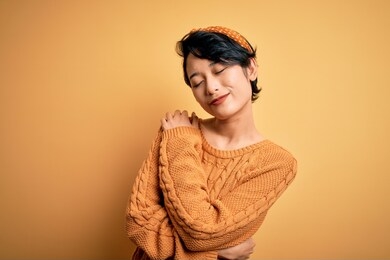 young beautiful asian girl wearing casual sweater and diadem standing over yellow background hugging oneself happy and positive, smiling confident. self love and self care