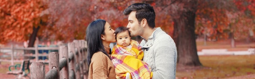 asian chinese mother and caucasian father dad holding kissing baby girl wrapped in blanket. family in autumn fall park outdoor. seasonal holiday thanksgiving. web banner header for website.