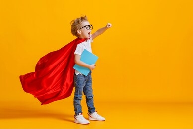 full body boy in superhero cape smiling and raising fist up while being ready for school studies against yellow backdrop