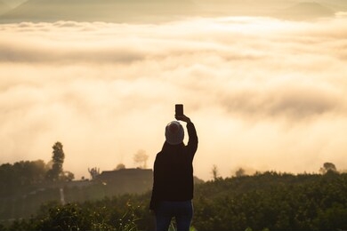 winter travel relax vacation concept, young traveler asian woman with sweater and wool hat taking photo and selfie with mobile phone on mountain with fog at sunrise in mae hong son, thailand