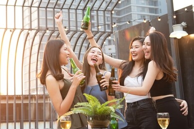 group of young beautiful happy asian women holding bottle of beer chat together with friends while celebrating dance party on outdoor rooftop nightclub with copy space for advertising.