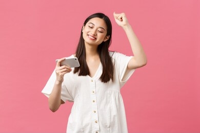 beauty, people emotions and technology concept. girl enjoying watching favorite k-pop music video on smartphone, dancing to beat, looking at mobile phone and chanting, pink background