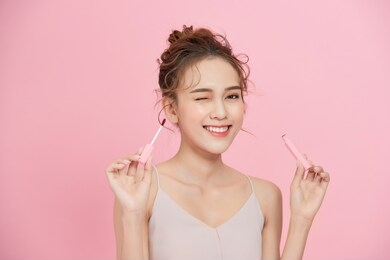 young smiling asian girl applying lipstick isolated over pink background.