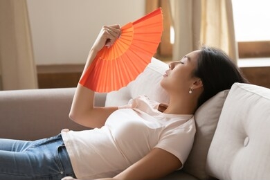 side view tired young asian ethnicity woman lying on sofa, using paper fan indoors, unhappy millennial vietnamese girl suffering from hot temperature at home, breathing fresh air, cooling herself.