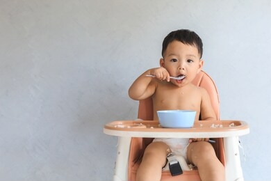 happy infant asian baby boy eating food by himself on baby high chair and making mess with copy space.