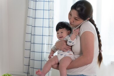 asian young mother working and holding baby while talking on phone in home office