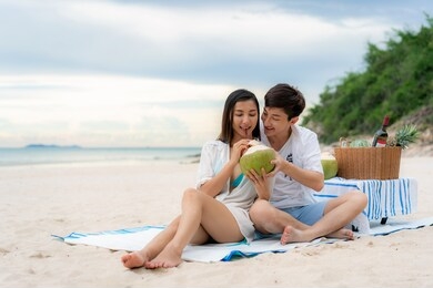 asian young couple sitting on the picnic blanket and drinking coconut water in the beach and near sea with tropical fruit in background. summer, holidays, vacation and happy people in thailand concept