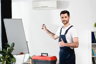 smiling worker holding remote controller and showing thumb up while standing near toolbox and air conditioner