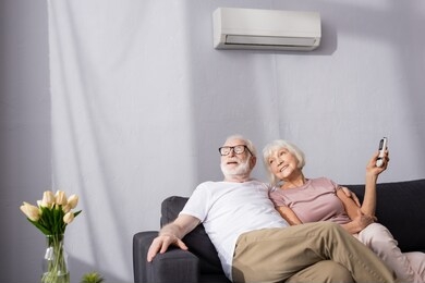 selective focus of smiling senior woman holding remote controller of air conditioner near husband at home