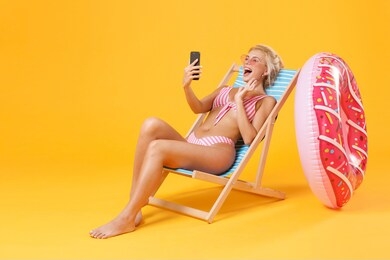 funny young woman in pink striped swimsuit glasses sit on deck chair isolated on yellow background. people summer vacation rest lifestyle concept. doing selfie shot on mobile phone, making video call