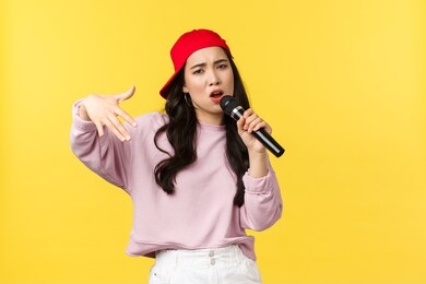 people emotions, lifestyle leisure and beauty concept. stylish and cool young girl rapper in red cap, singing song and gesturing, performing with microphone, standing yellow background
