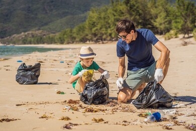 dad and son in gloves cleaning up the beach pick up plastic bags that pollute sea. natural education of children. problem of spilled rubbish trash garbage on the beach sand caused by man-made