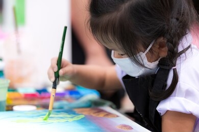 pupils girl are concentrating on drawing and painting with brush and watercolor on the canvas. an asian little child is wearing a white cloth face mask while learning to paint. child 3 years old.