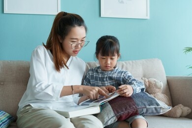 asian family with mother and daughter reading book together while sitting on the sofa in the living room at home morning