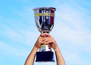 first price to winner, cup trophy in silver and gold. hands holding it up, cloudy but blue sky. 