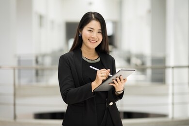 portrait of a young cheerful businesswoman surfing social network on digital tablet in front of office during break. asian business woman standing in office building. ฺbusiness stock photo.