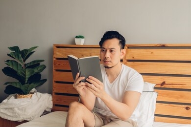 asian man is reading book on his bed in late afternoon.