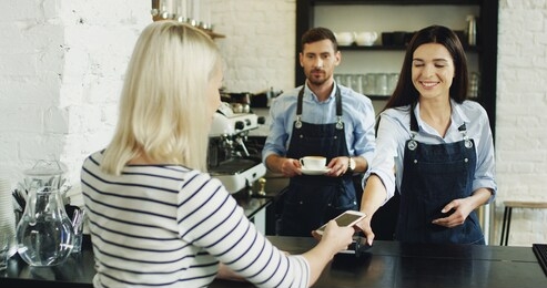 rear of the blond woman paying with her smartphone at the bar while buying a coffee and waiter serving it. in the cafe.