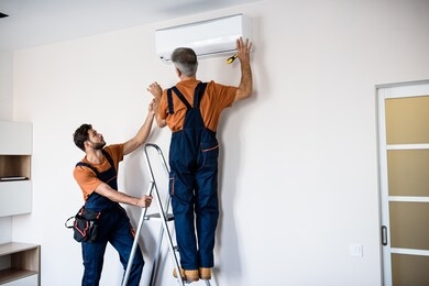 two workers in uniform, air conditioning masters using ladder while installing a new air conditioner in the apartment. construction, maintenance and repair concept. horizontal shot